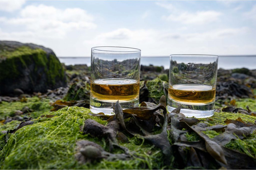 Two glasses of whisky sit on a Scottish coast