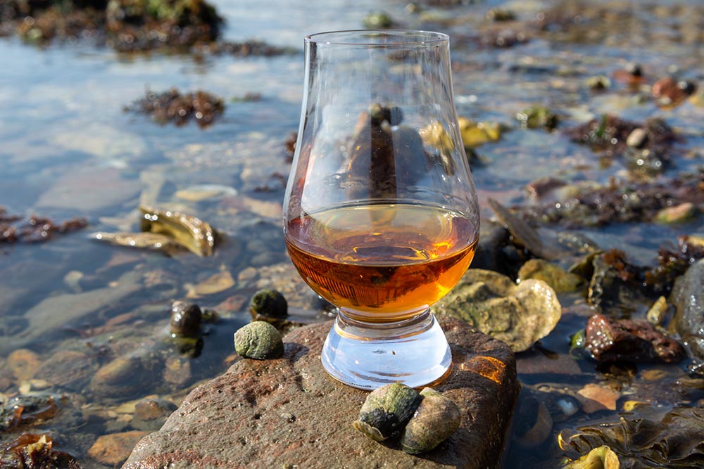 A dram of whisky sits on a rock in the sea.